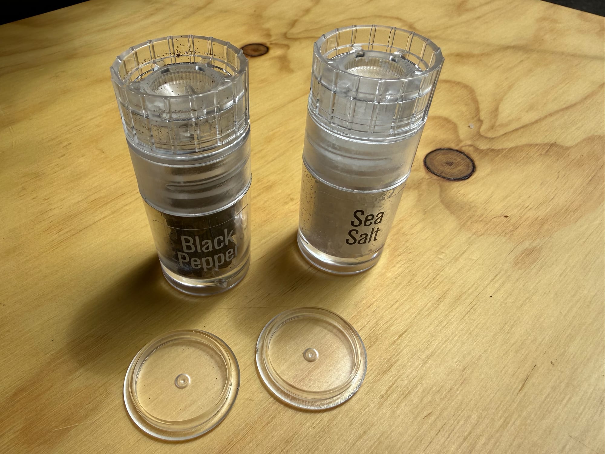 The AGS mini black pepper and sea salt grinders on a wooden table with their clear caps in front