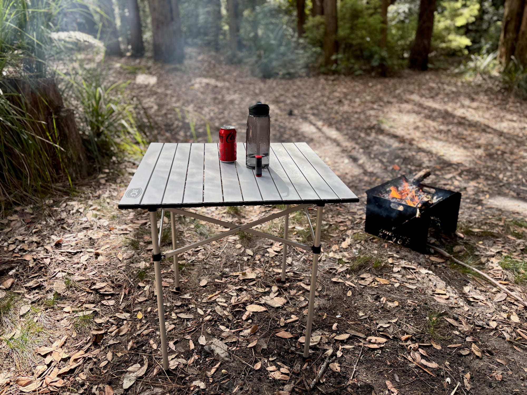 BCF aluminium roll up camp table set up at a bush campsite, with a drink and a bottle on top