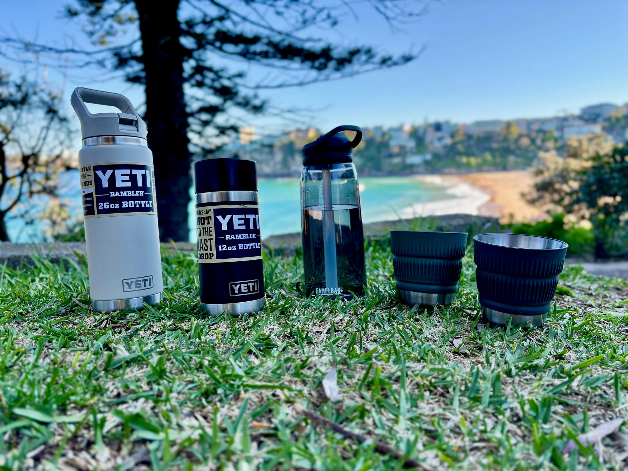 Two YETI Rambler bottles, a CamelBak Eddy+ and two insulated cups lined up on the grass above a beach