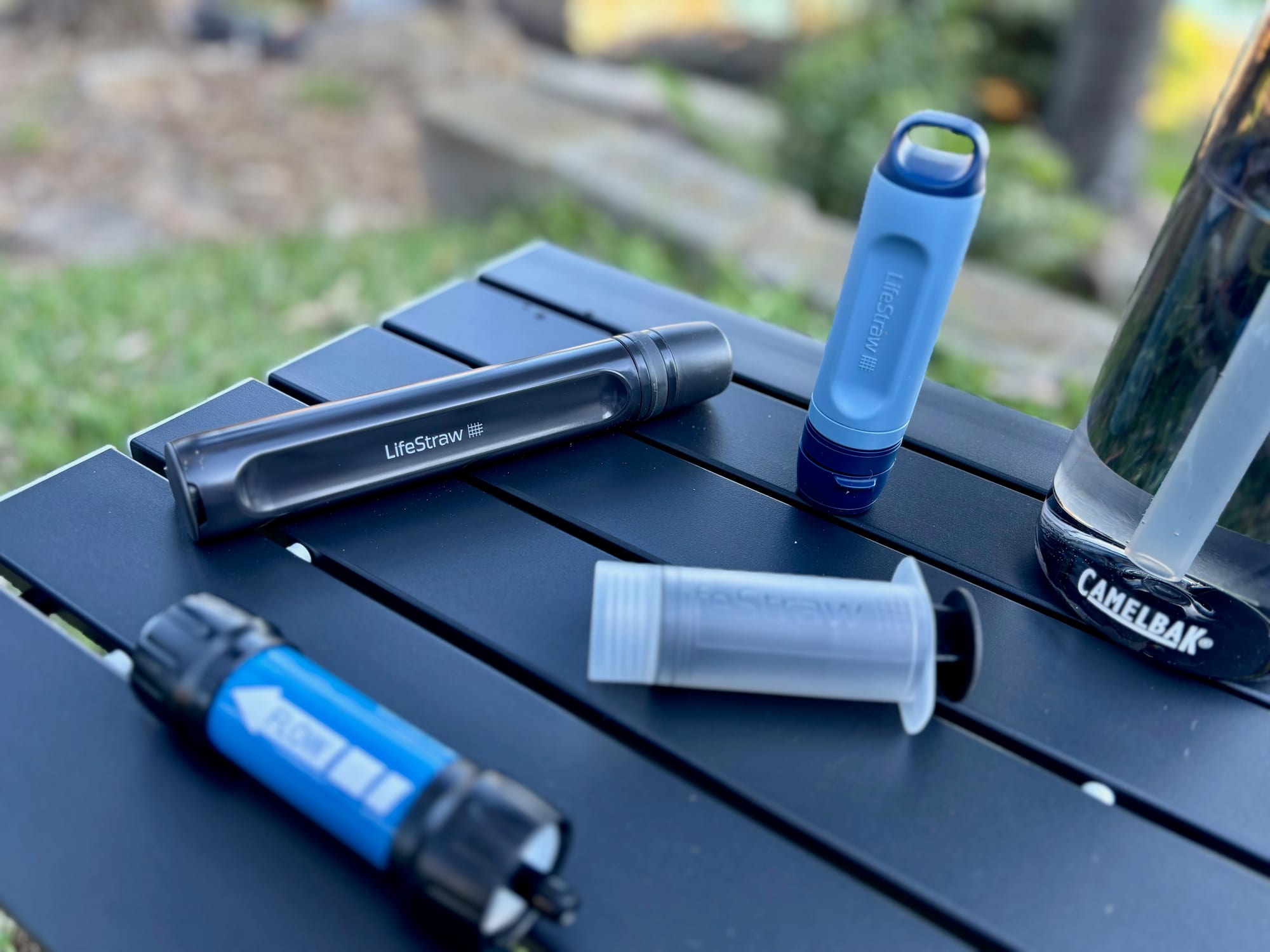 Four water filters and a backwash syringe on a camp table, with a water bottle behind and the sea in the distance