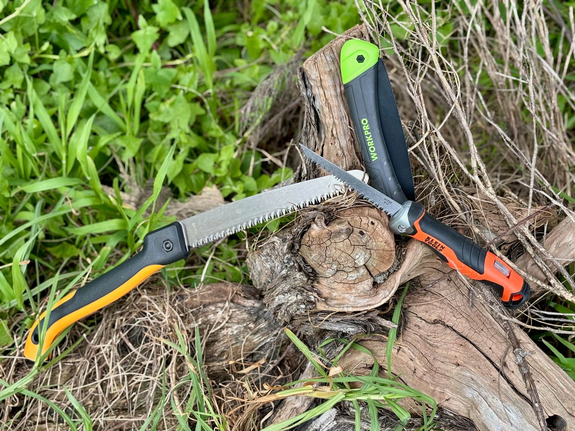 Folding saws with the blades open on a tree stump: a Fiskars, a Workpro and a Klein Tools