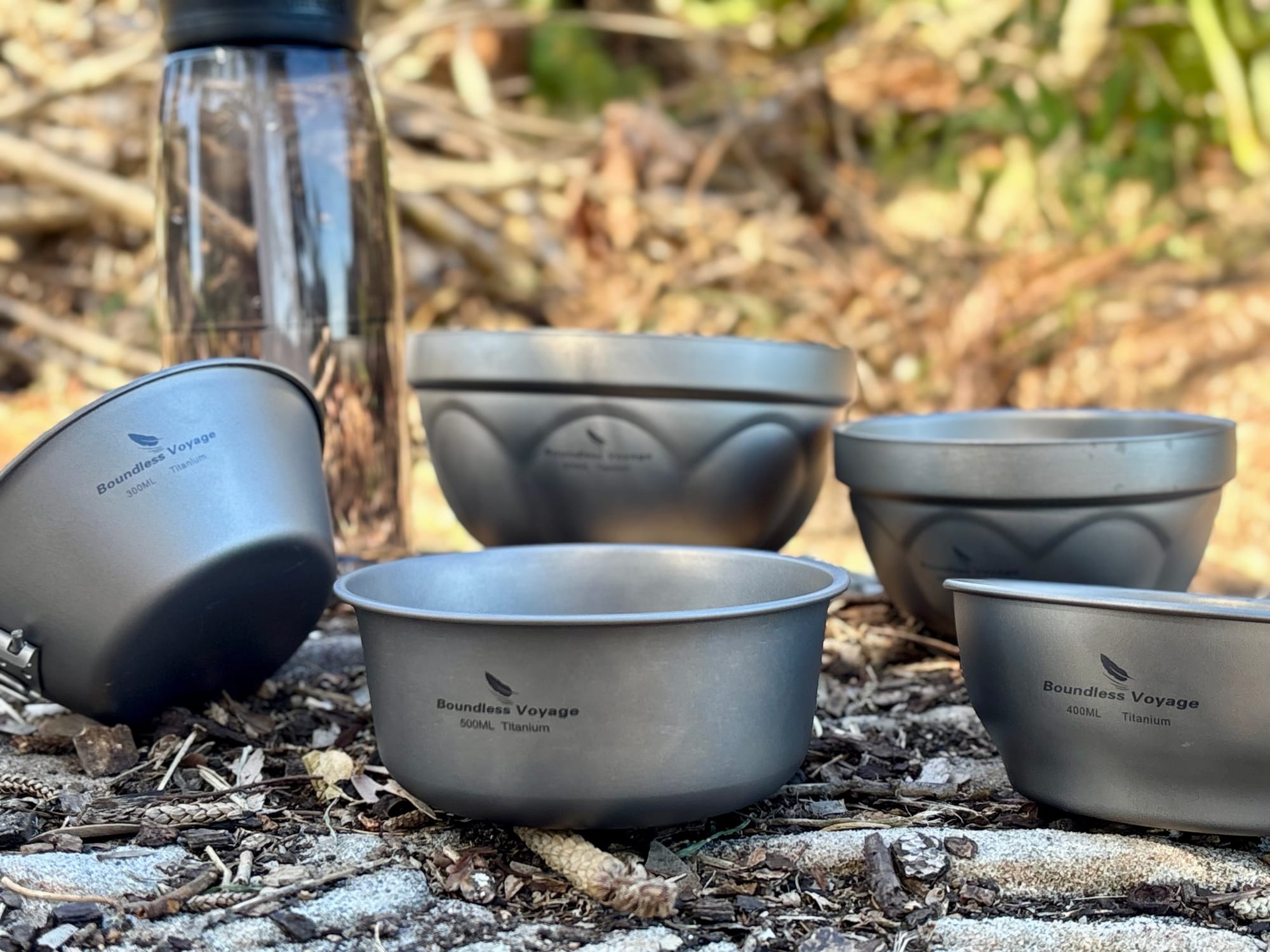Boundless Voyage titanium bowls and a bowl with a folding handle on a rock, with a drink bottle behind