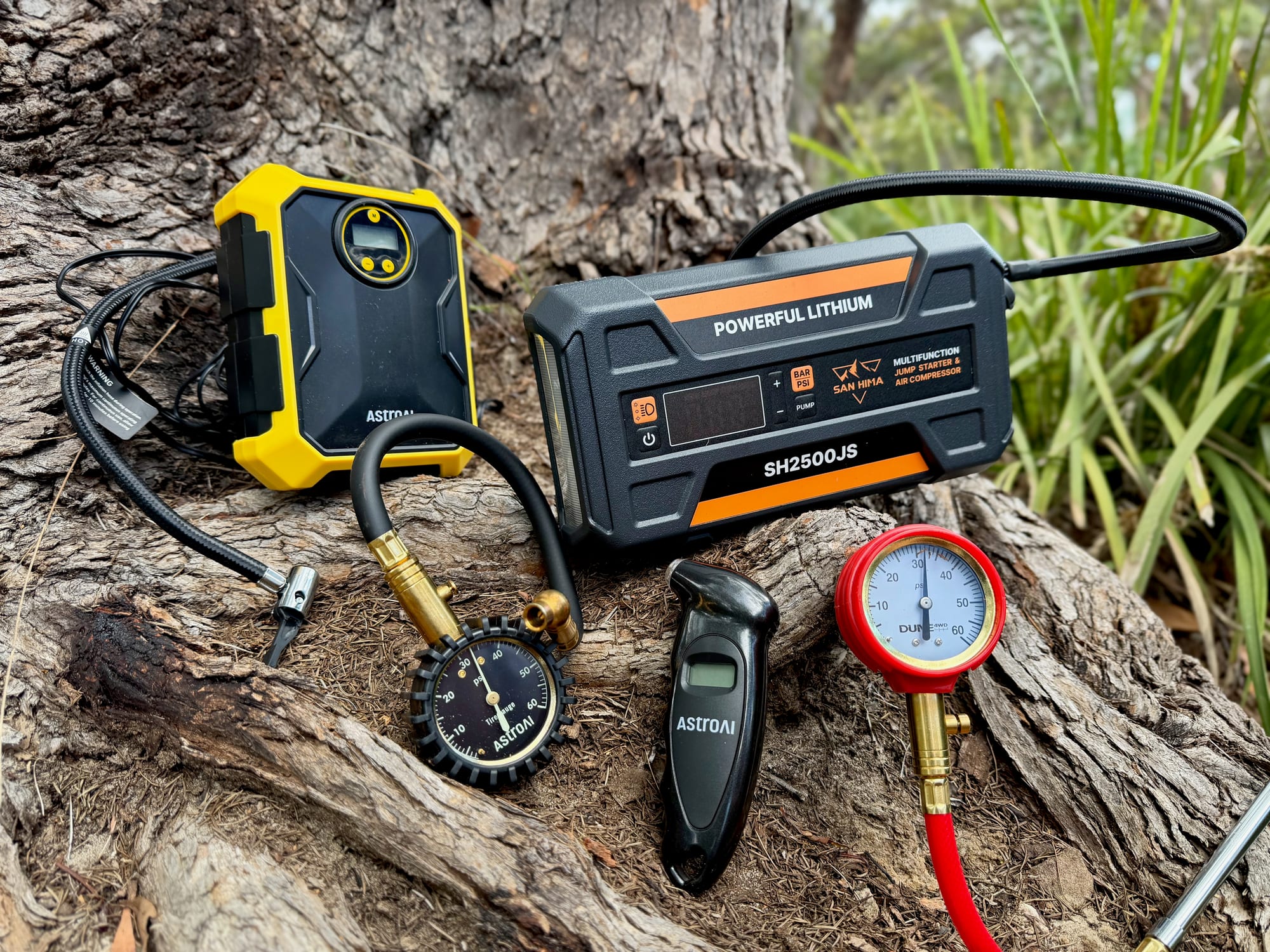 The AstroAI tyre inflator, a San Hima jump starter with compressor and three tyre pressure gauges on a tree root