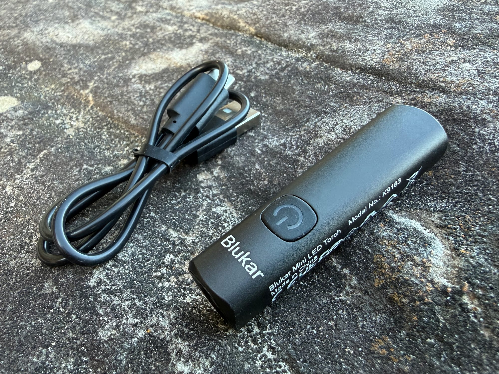The Blukar Mini Torch next to its USB-C charging cable on a rock