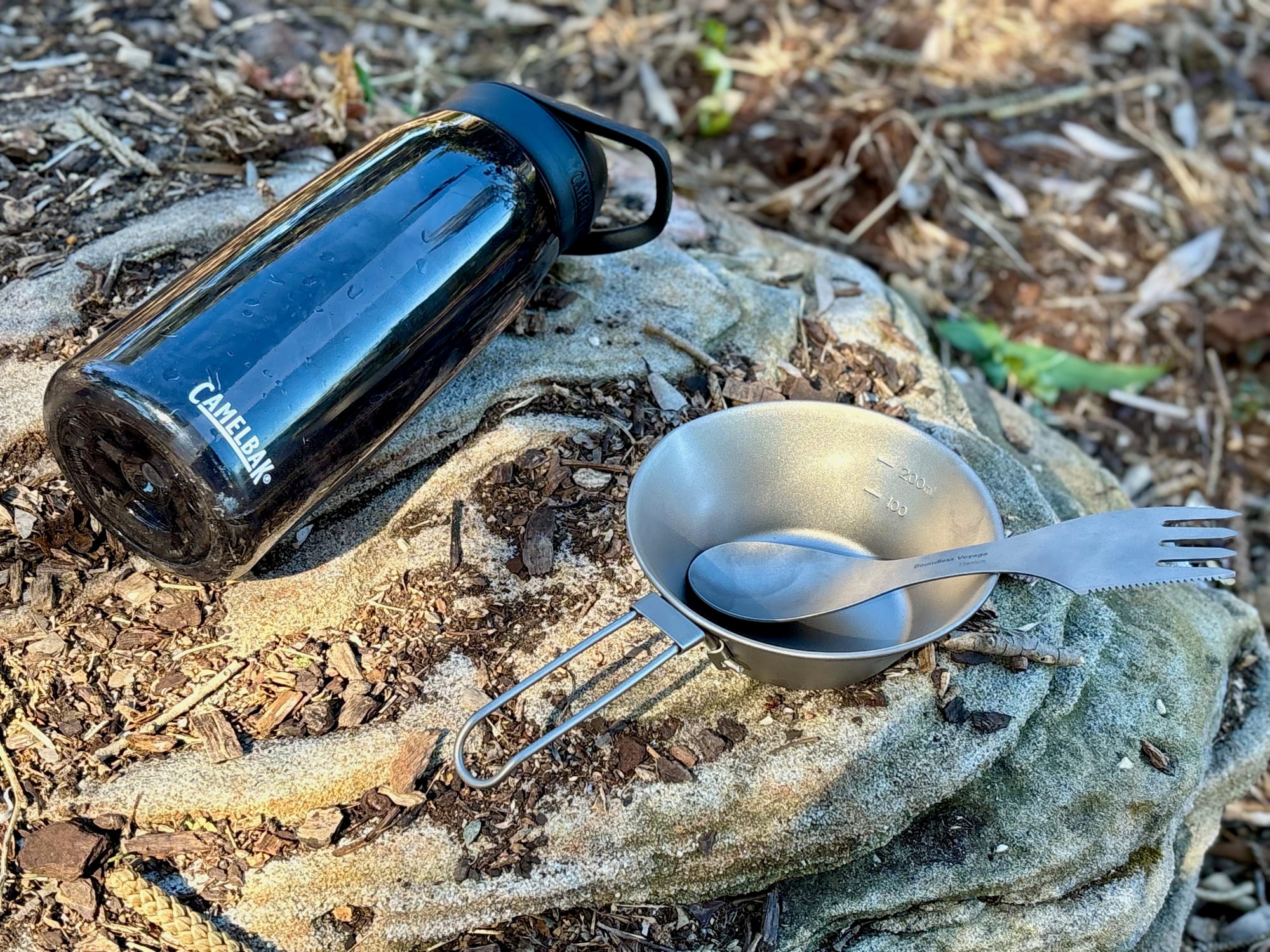 The 300 ml titanium bowl with a spork in it next to a CamelBak drink bottle lying on a rock