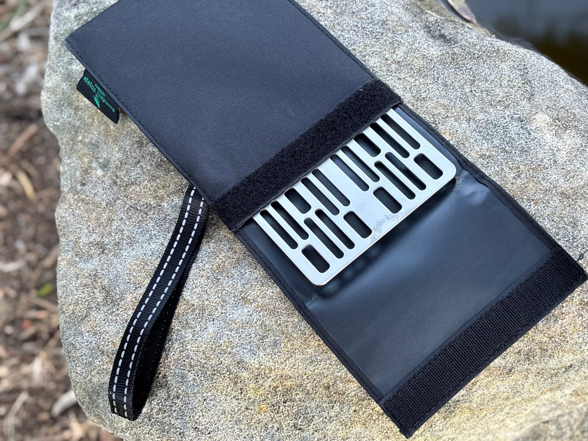 The Ti2023C titanium grill plate tucked into its black sleeve on a rock, with the carry loop beside it