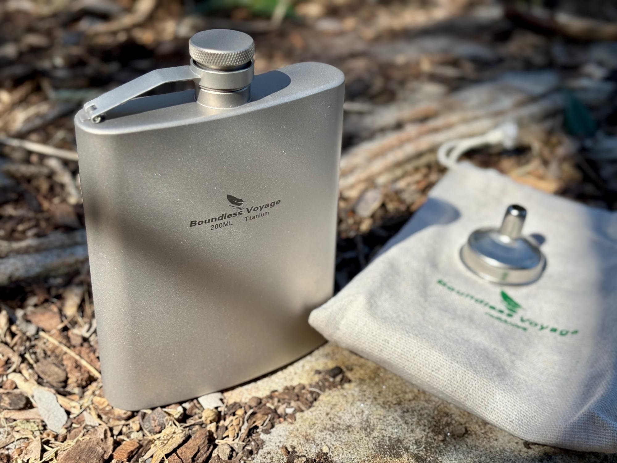 The Boundless Voyage 200 ml titanium hip flask with its funnel and cotton pouch, on bark chips