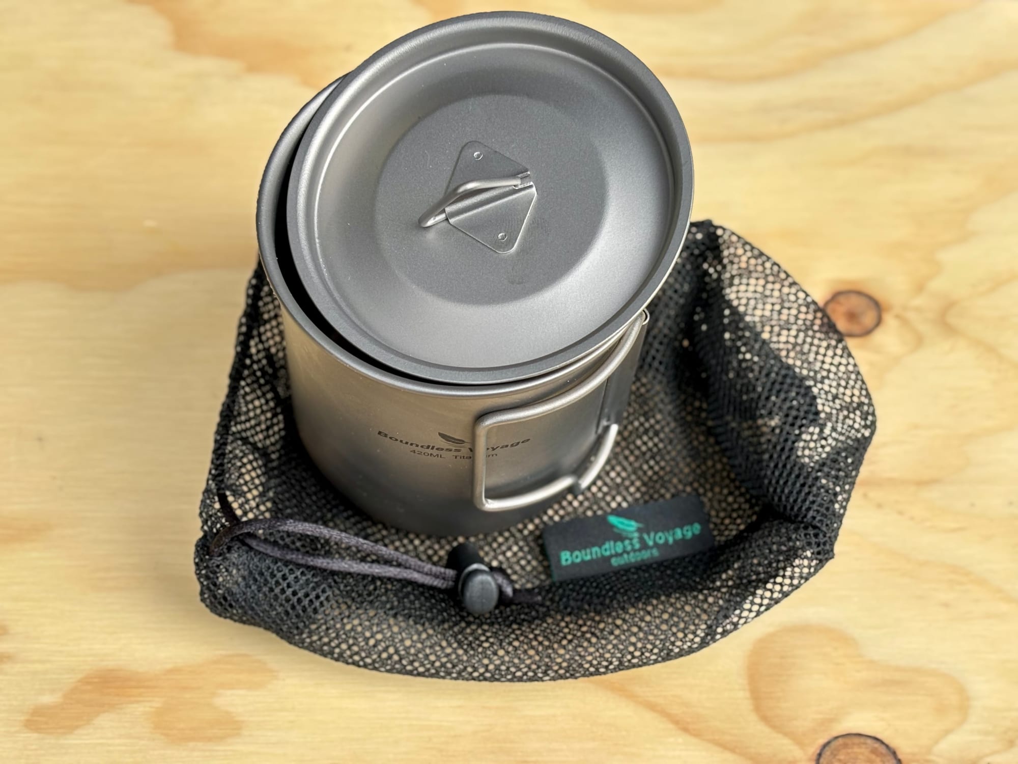 The titanium mug with its lid on, sitting in its black mesh bag with a Boundless Voyage tag