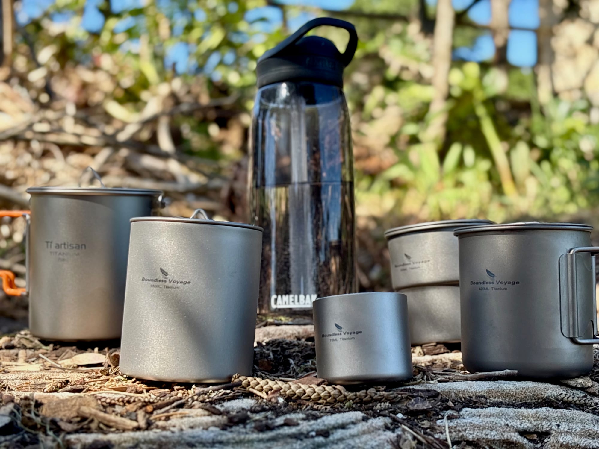 Boundless Voyage and Ti Artisan titanium mugs and cups lined up next to a CamelBak bottle