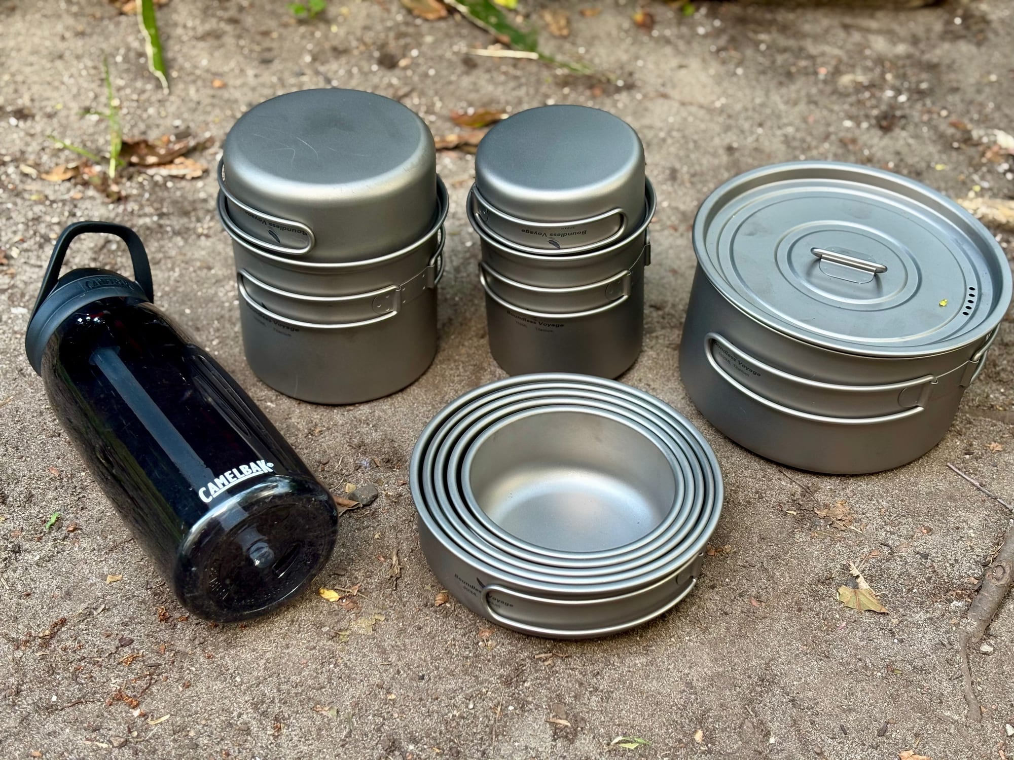 The nested Boundless Voyage pan set, the 750 + 300 ml and 1000 + 500 ml pot sets, a larger lidded titanium pot and a 1 litre CamelBak bottle on sand