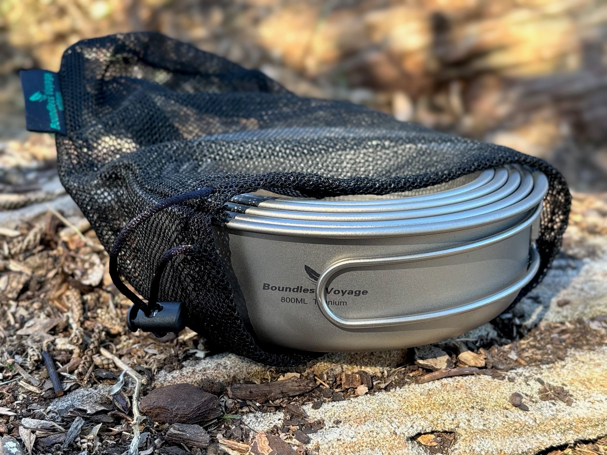 The nested Boundless Voyage titanium pans half inside their black mesh bag, seen from the side on a rock