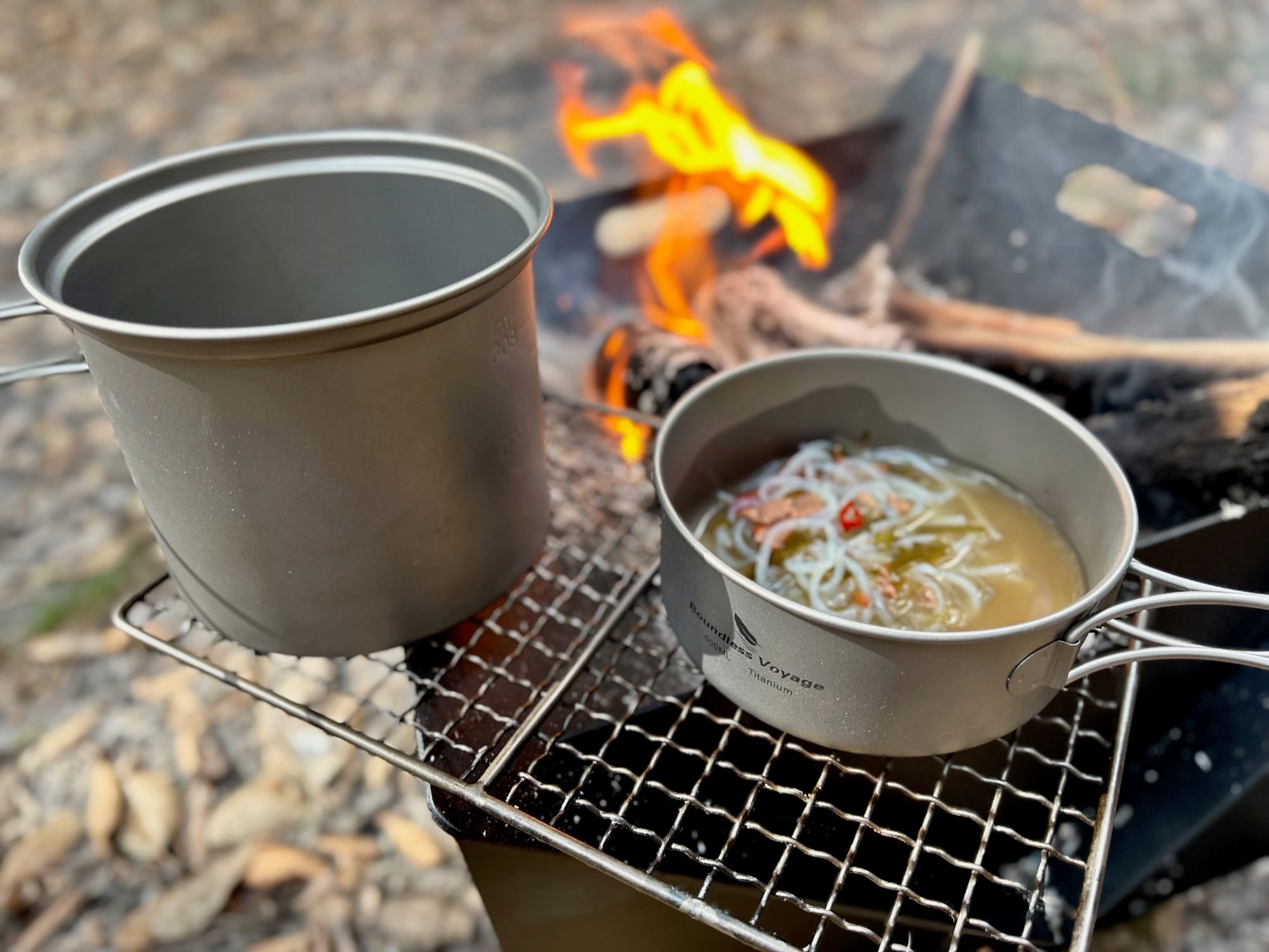 The titanium pot and the pan side by side on a grill over the fire