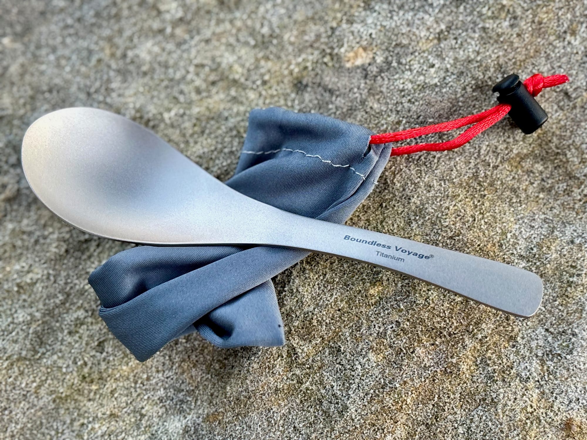 The Boundless Voyage titanium spoon on its grey drawstring pouch on a sandstone rock