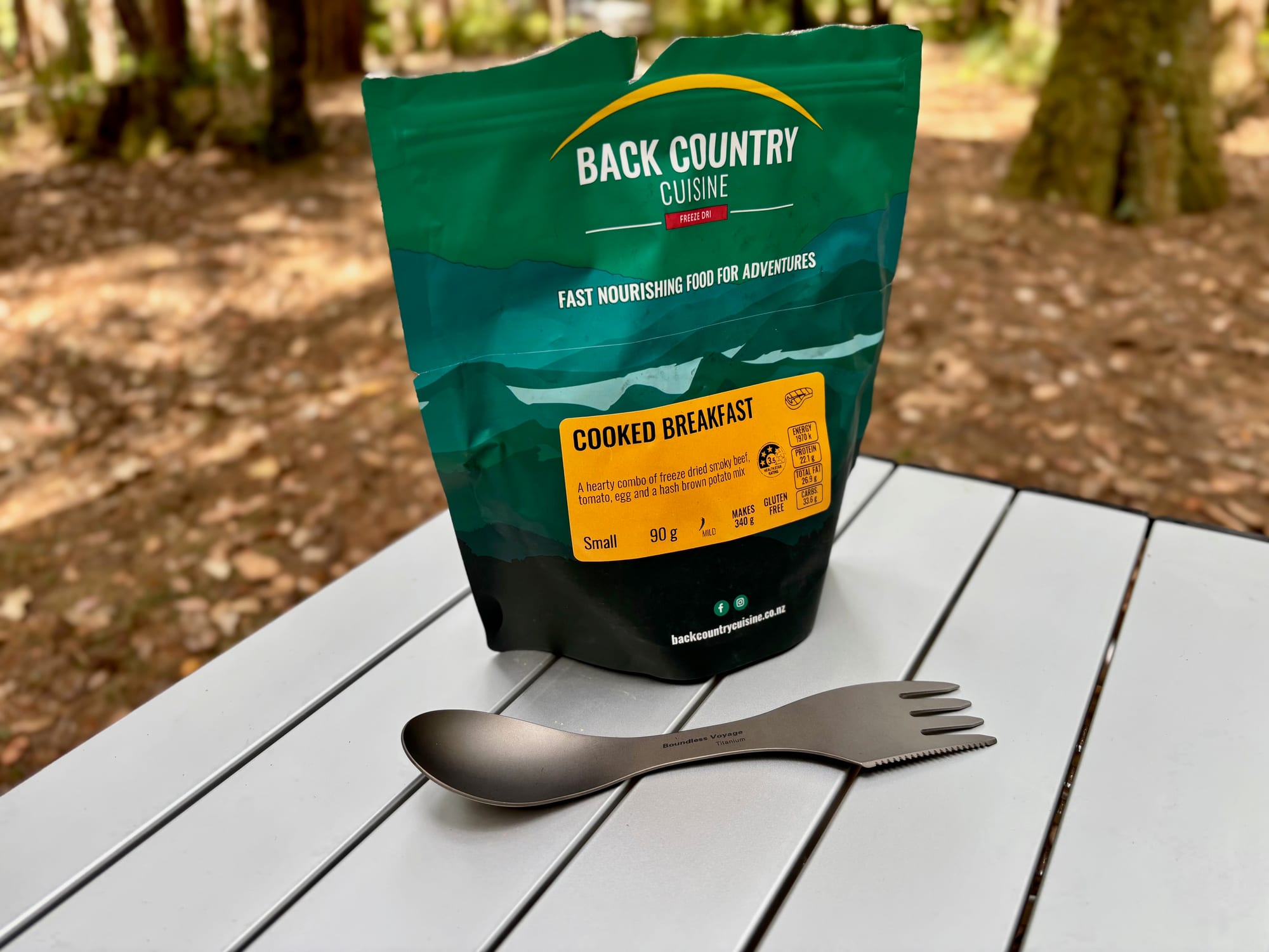 The Boundless Voyage titanium spork lying in front of a Back Country Cuisine meal pouch, showing the spoon and fork ends