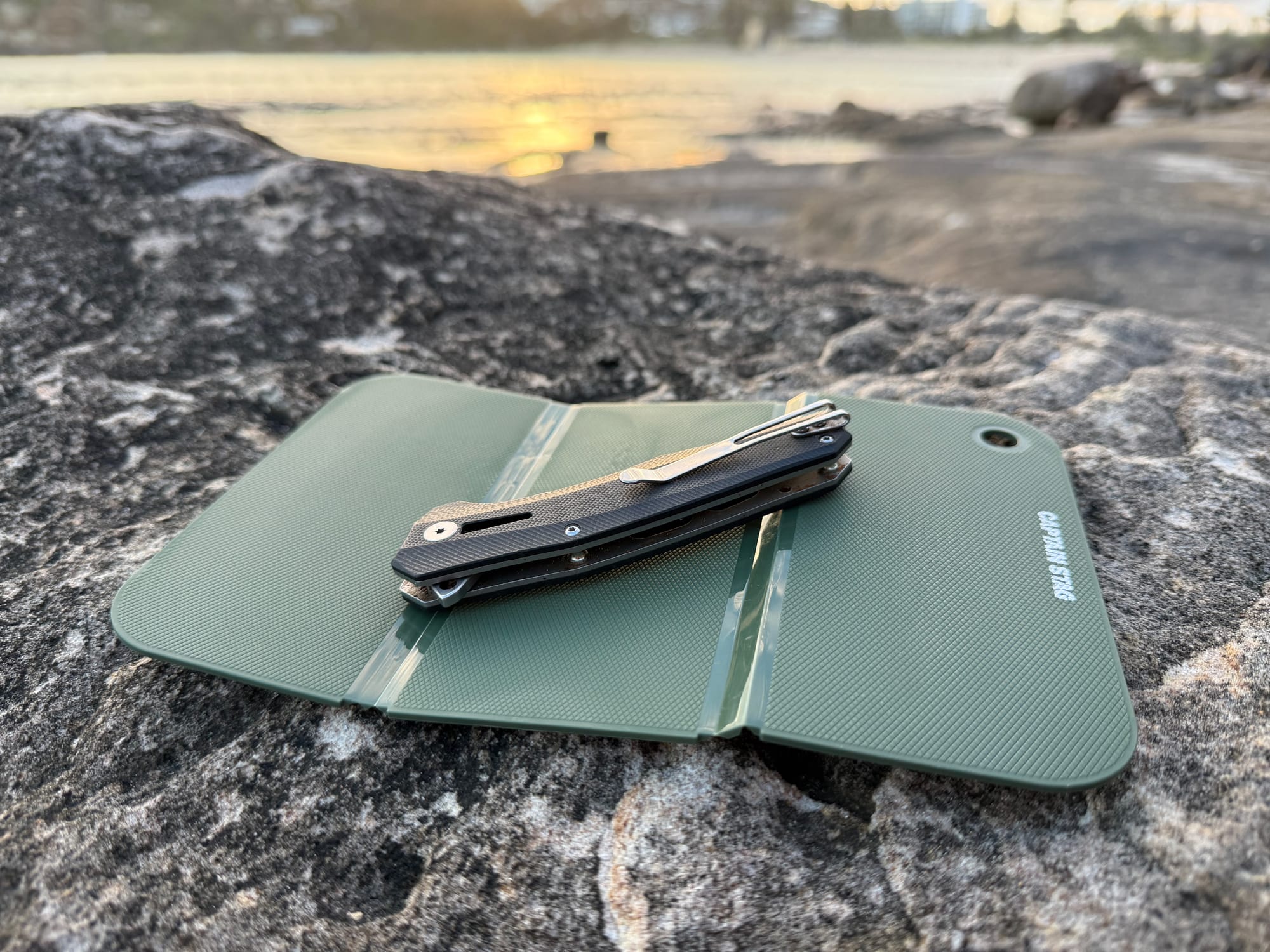 The Captain Stag cutting board with a folding knife on a rock by the water at sunset