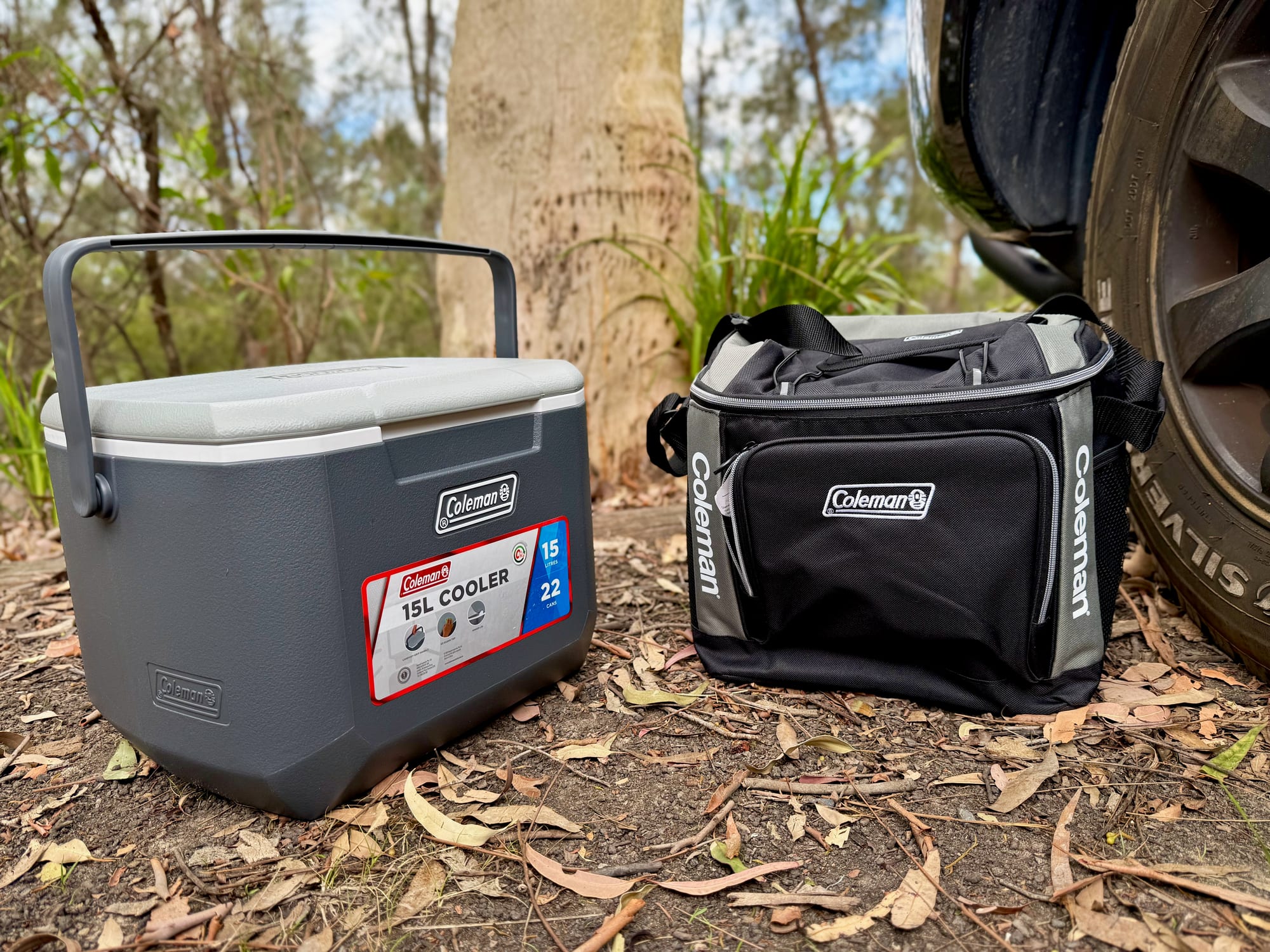 The Coleman 15L hard cooler and the Coleman 30 Can Soft Cooler side by side on the ground at camp