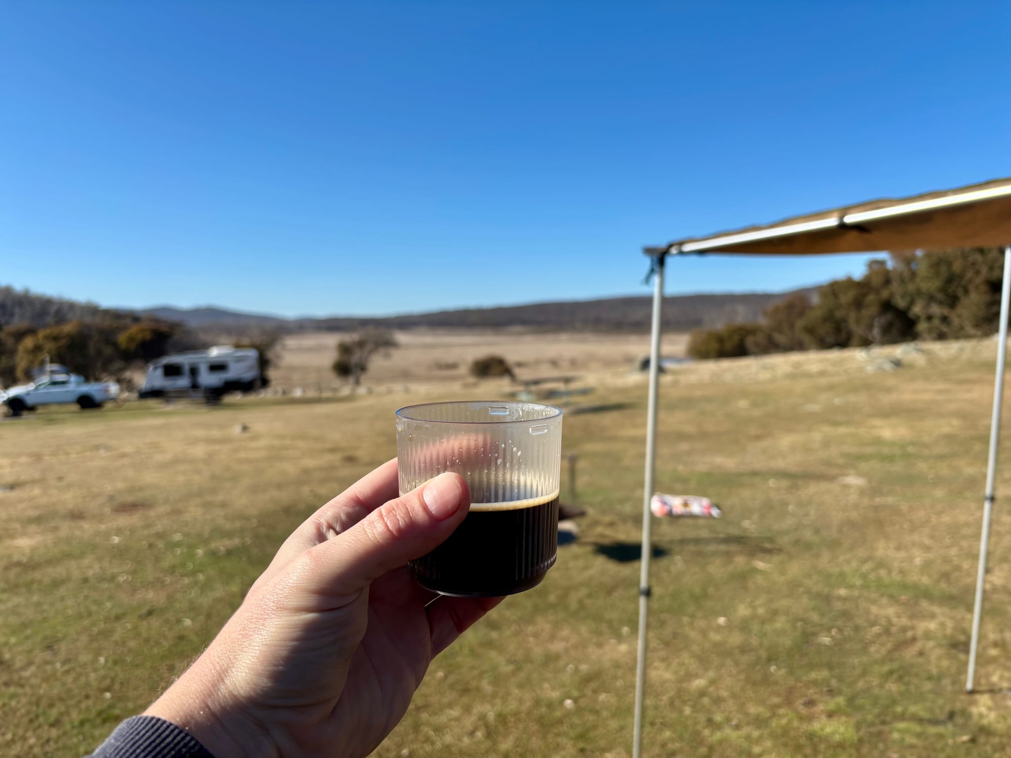 A cup of coffee held up in front of the awning, with the open plain of Denison Campground behind