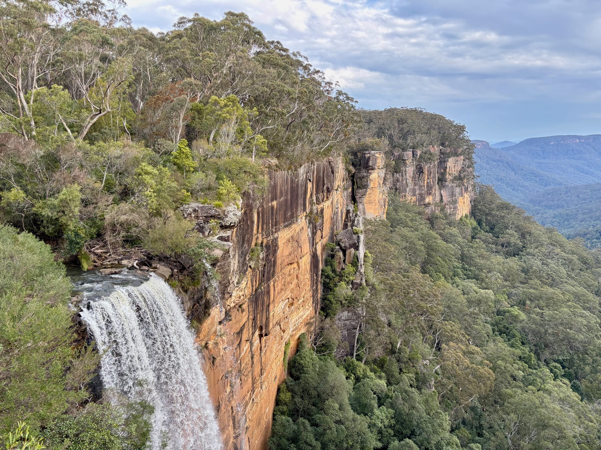 The top of Fitzroy Falls going over the orange sandstone cliff, with the valley beyond