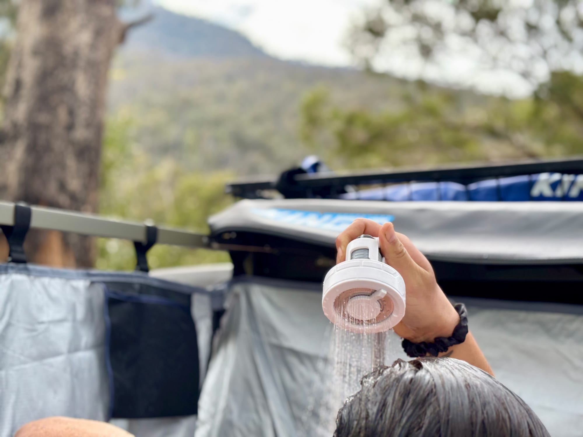 The FlexTail Max Shower held overhead and spraying onto wet hair at camp, with the shower tent and forested hills behind