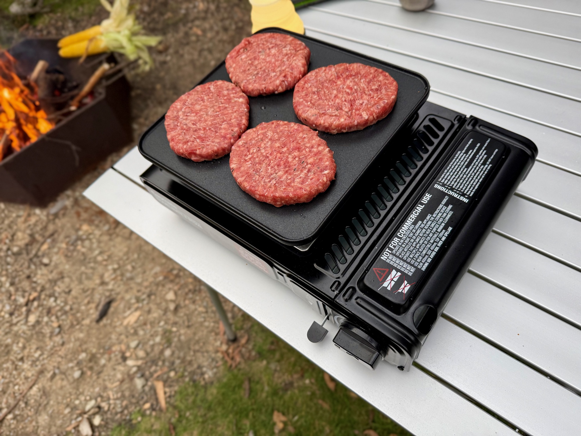 Four burger patties on the Gasmate Travelmate II hotplate, with a campfire behind
