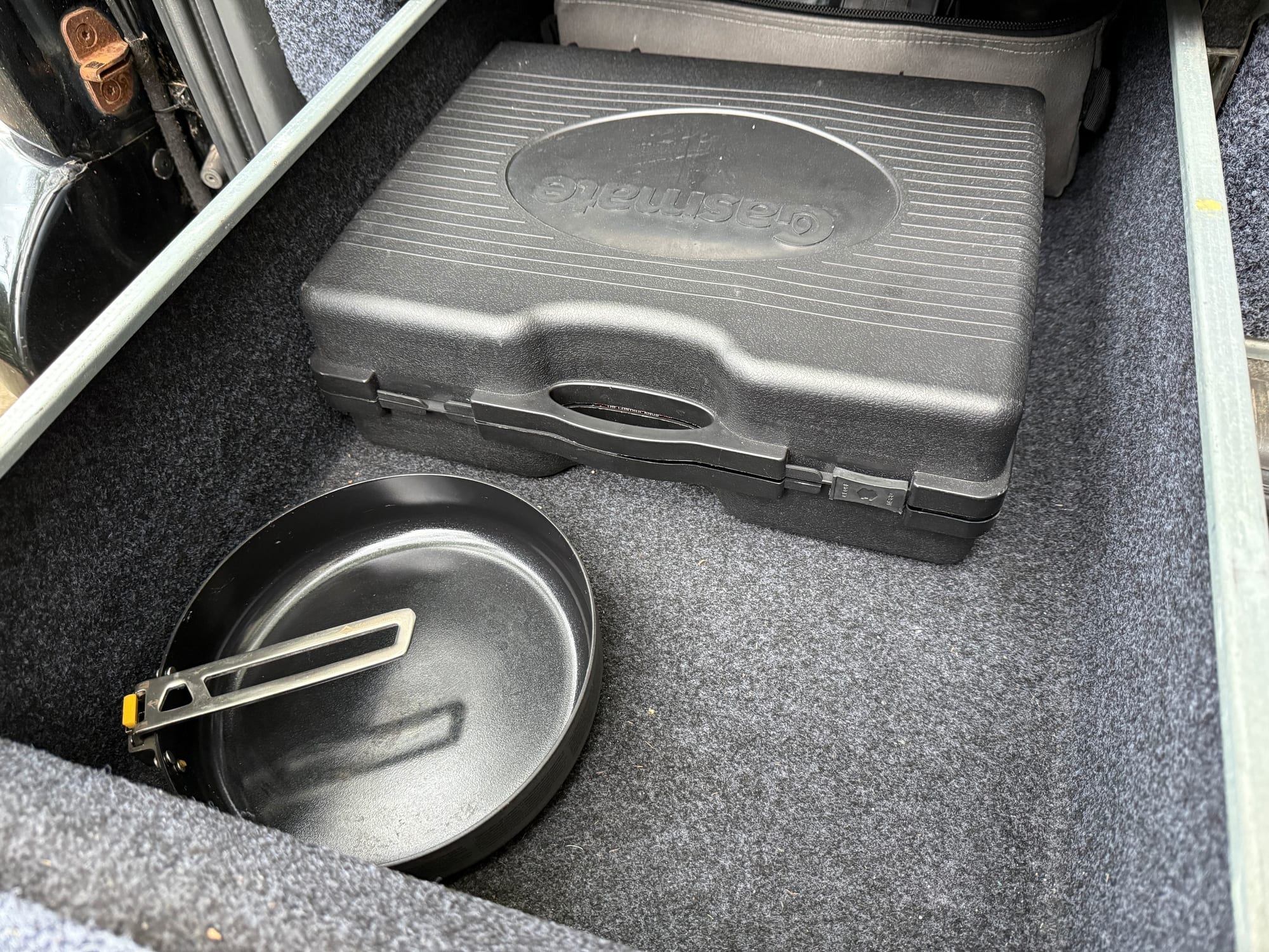 The Gasmate Travelmate II in its black carry case in a carpeted 4WD drawer, next to a frying pan