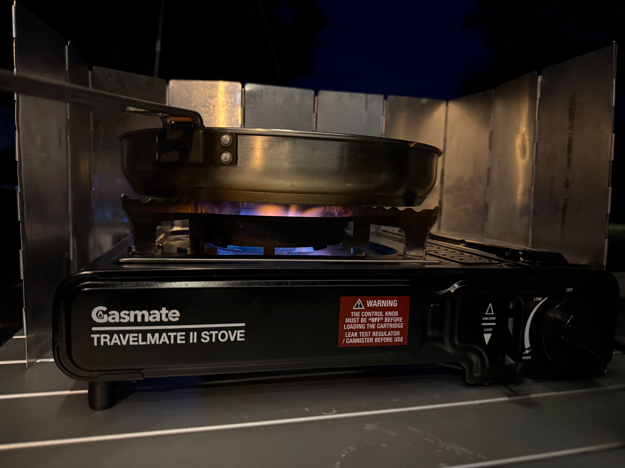 The Gasmate Travelmate II burner lit behind a windshield at night, with a frying pan on top