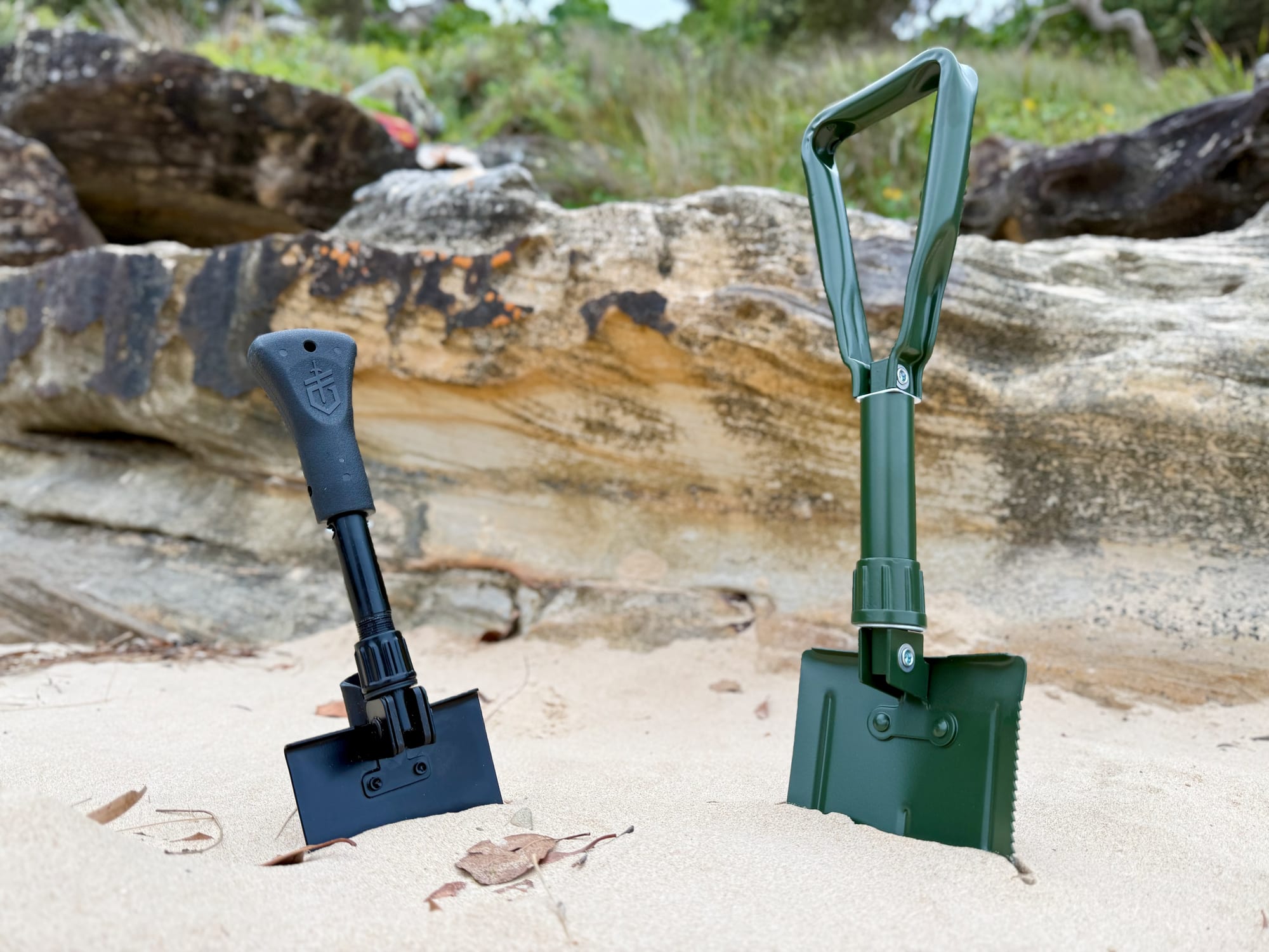 The black Gerber Gorge folding shovel standing in the sand next to a taller green folding shovel