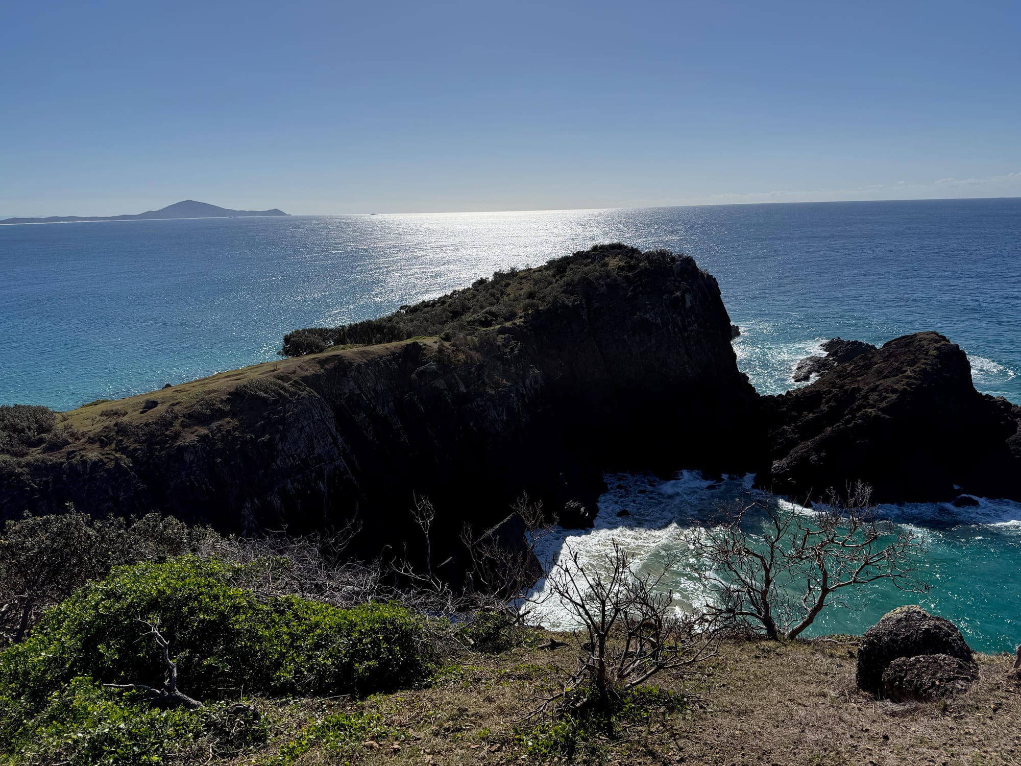 A dark rocky headland with waves breaking around it and the sun on the sea behind, from the Korogoro walking track