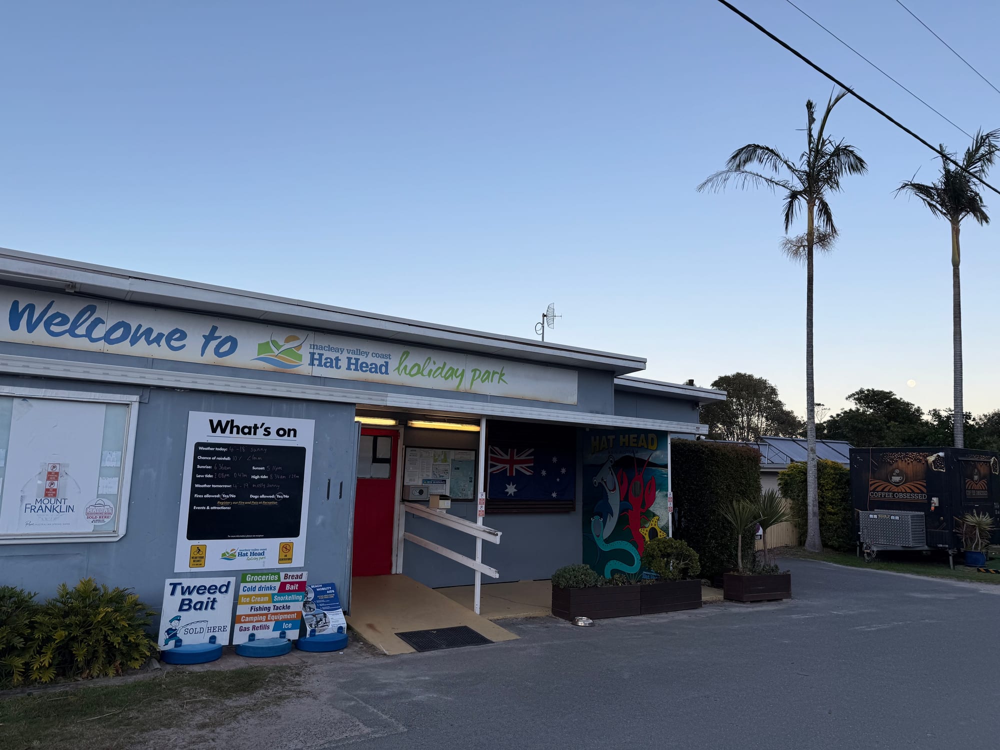 The reception building of Hat Head Holiday Park, with a What’s on board and palm trees out the front