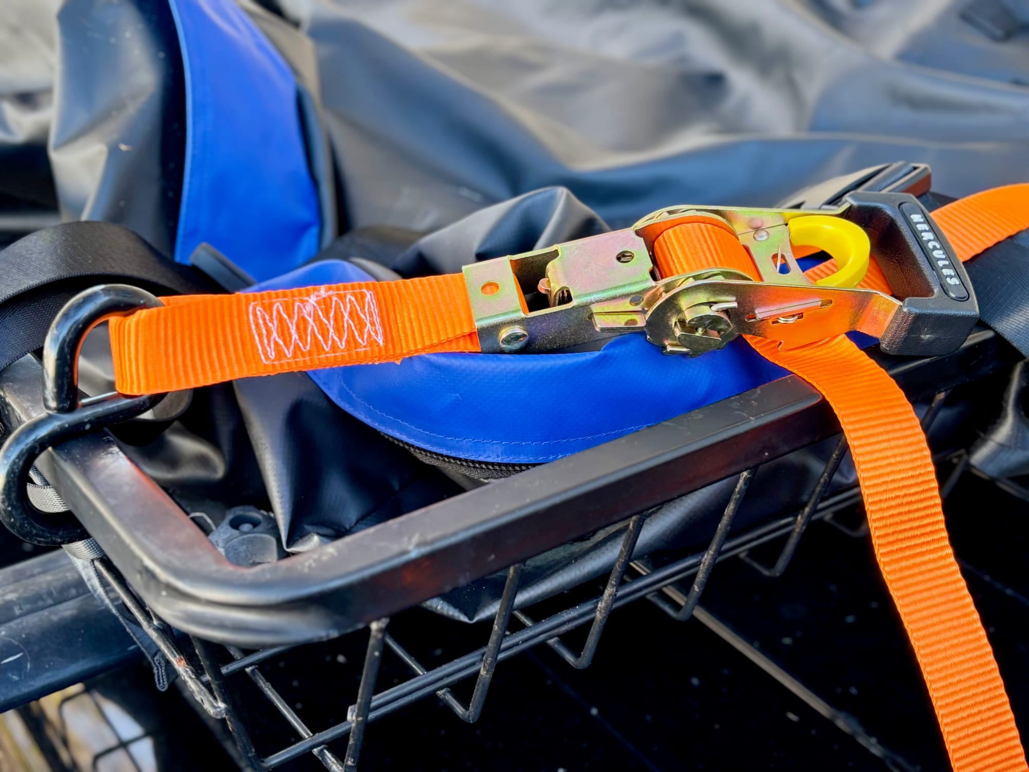 A Hercules ratchet strap holding a bag down in a roof basket, with its hook on the basket’s bar