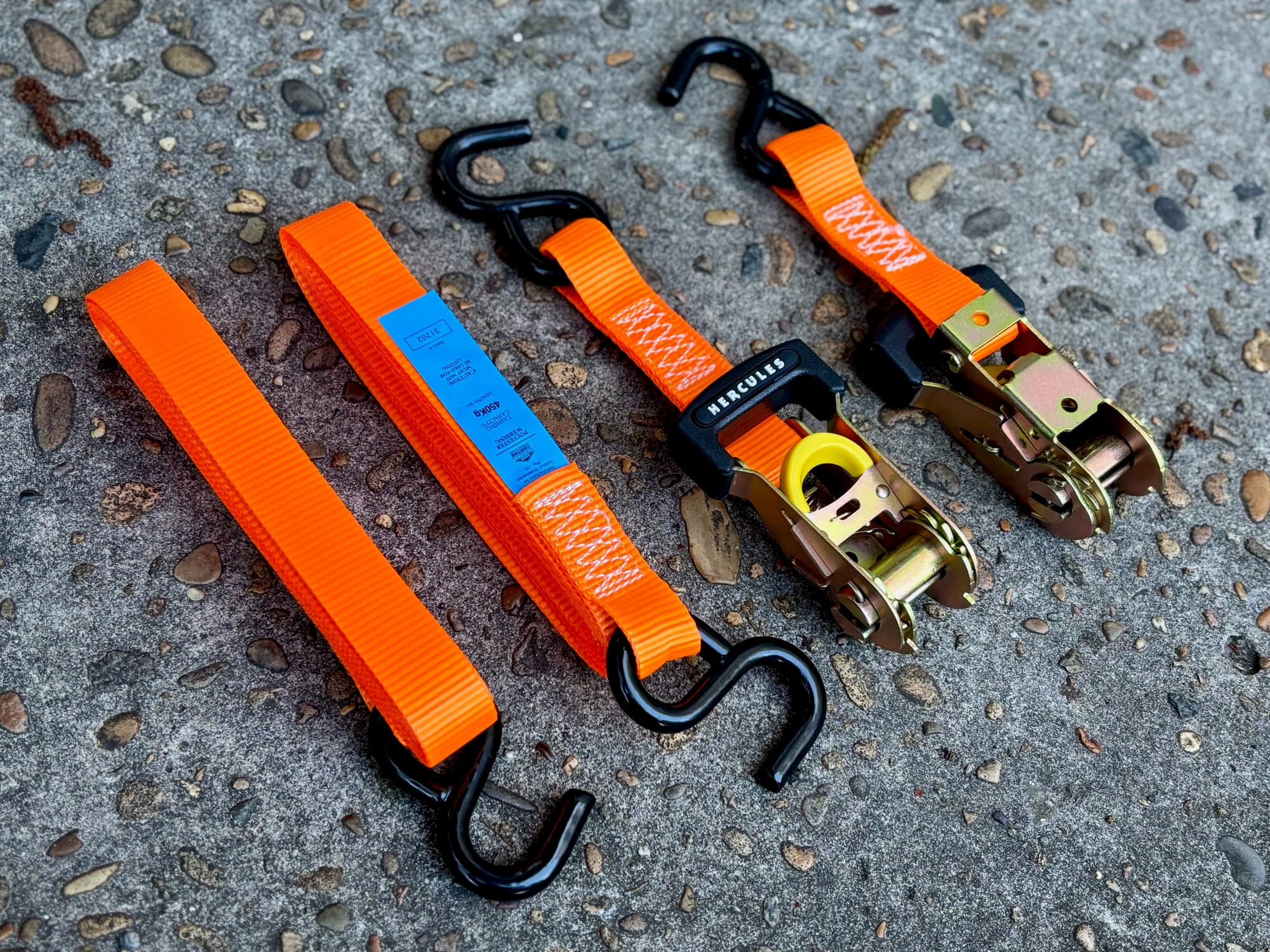 Two orange Hercules ratchet straps laid out on the ground, each with a ratchet, a long strap and black S-hooks