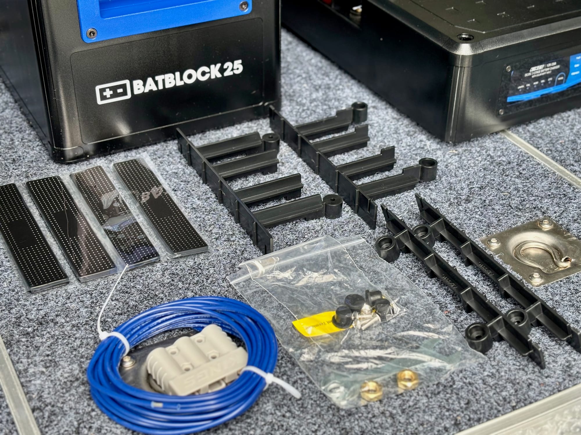 The parts from the BatBlock 25 box laid out: four black foam pads, black plastic spacers, the blue wire with a grey Anderson-style plug and a bag of bolts and knobs