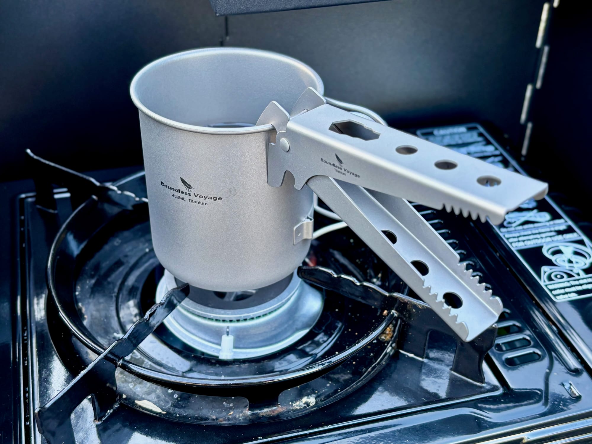 A titanium pot gripper clamped onto the rim of a titanium cup of hot water on a butane stove