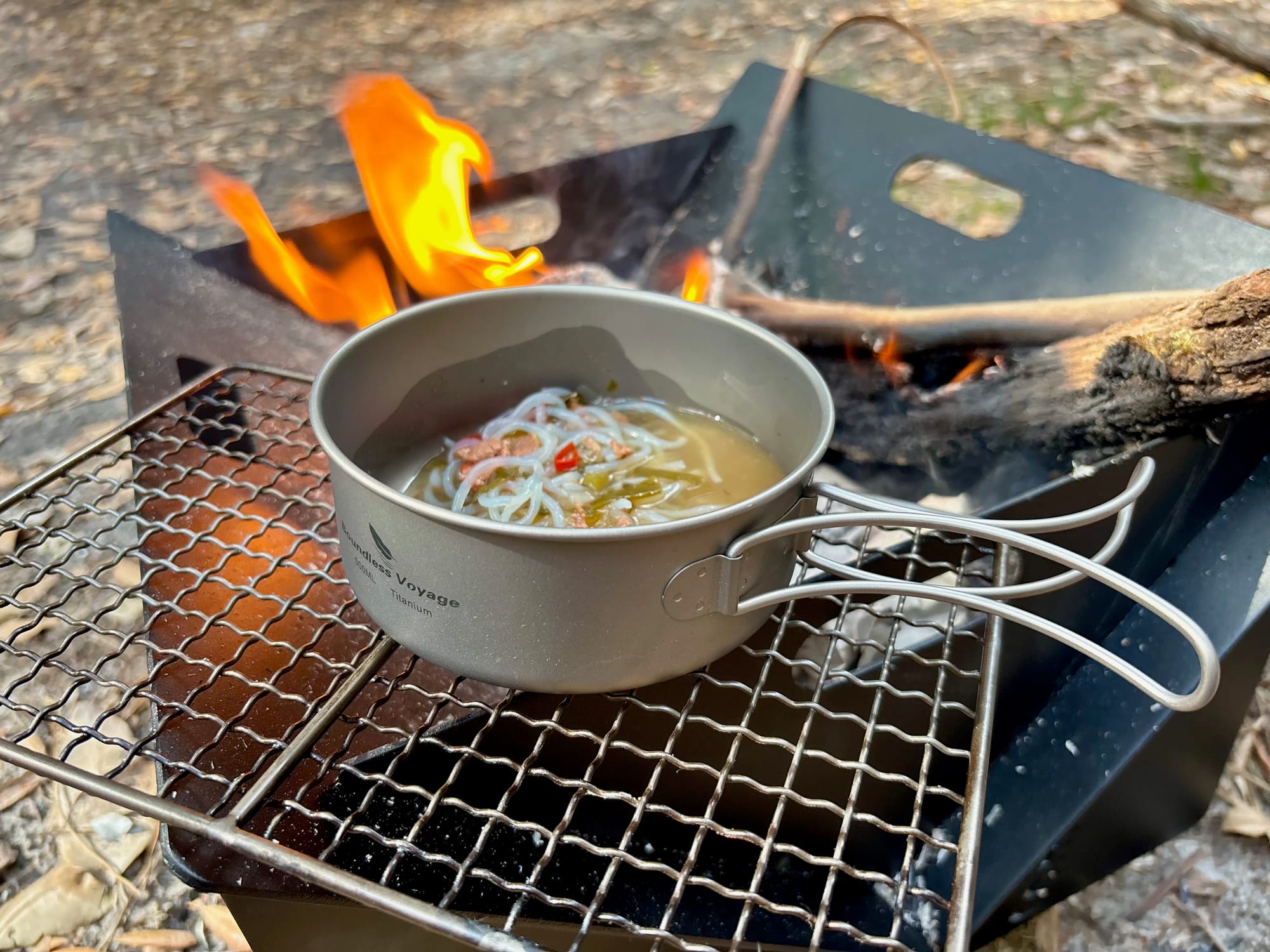 A titanium pan of noodle soup cooking on a grill over a fire pit, with flames behind