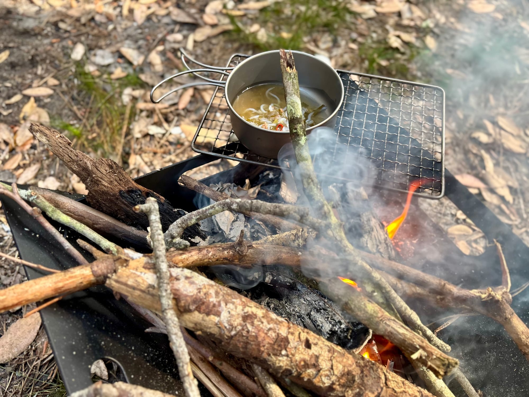 A titanium pan on a grill over a wood fire, with smoke rising
