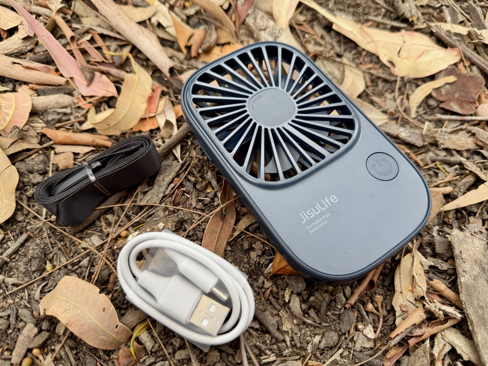 The navy JisuLife Life2 on dry leaves next to its neck strap and USB-C cable
