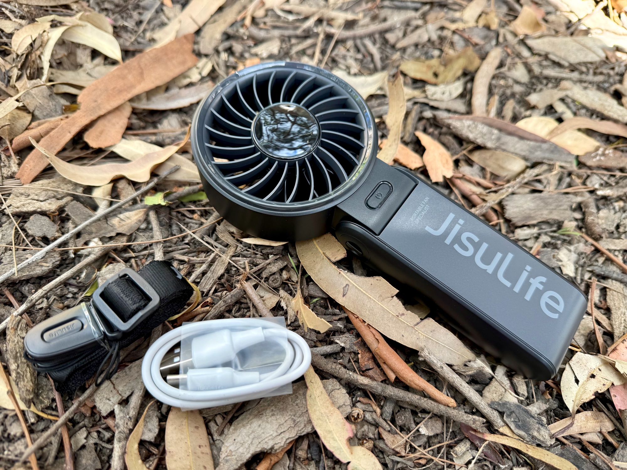 The JisuLife Life7 with its lanyard and USB-C charging cable on leaf litter