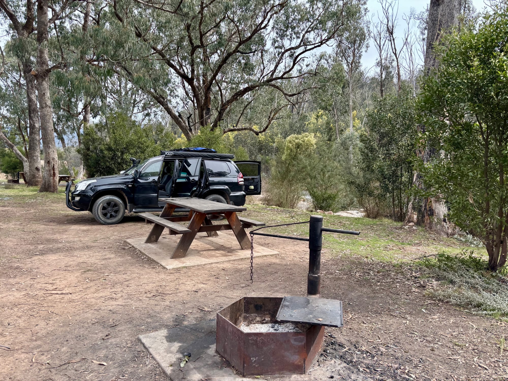 A steel fire pit with a swing-out hotplate at our site, with the picnic table and our 4WD behind