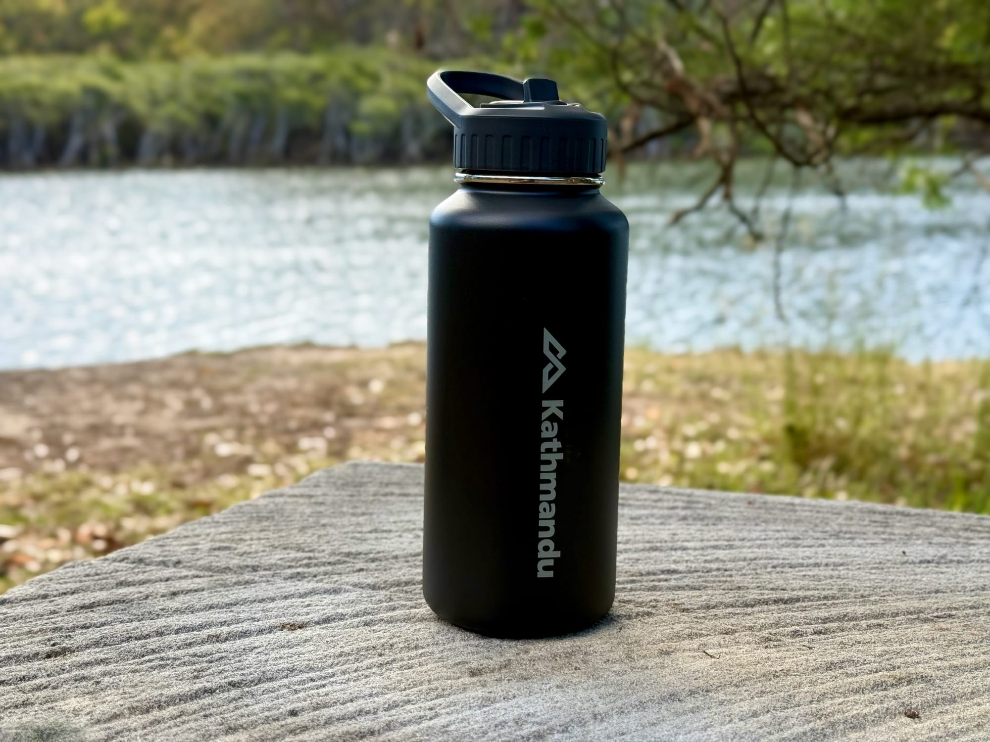 The Kathmandu Straw Lid Insulated Bottle standing on a rock by a lake
