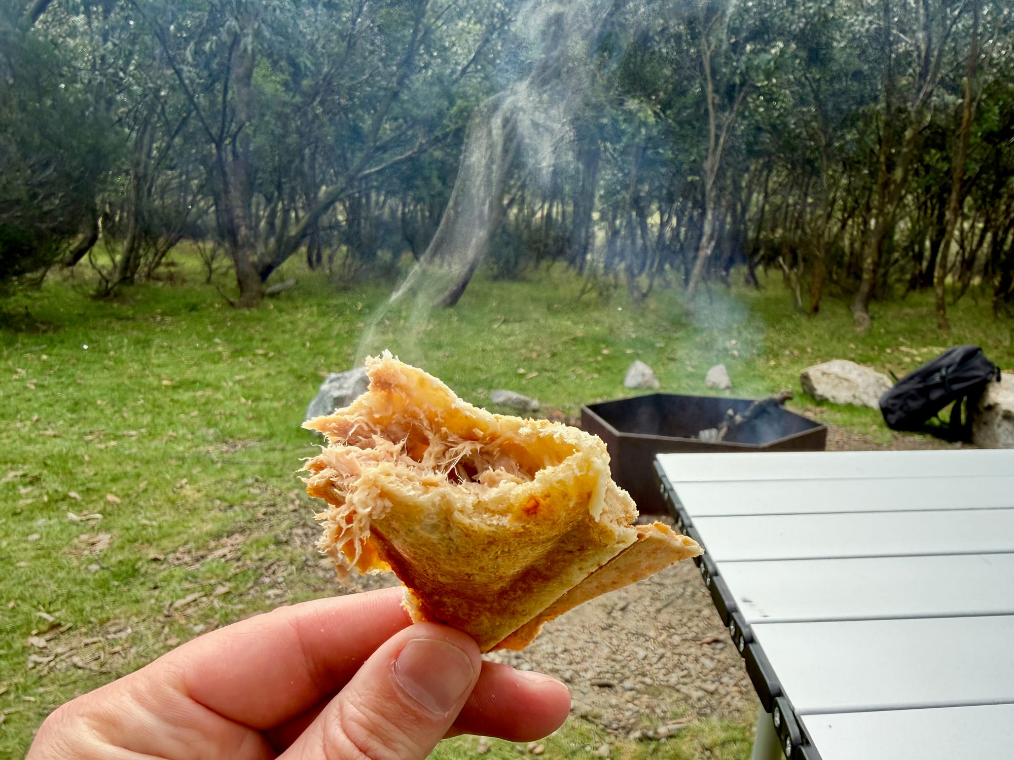A jaffle with melted tuna and cheese, held up at camp with the fire pit behind