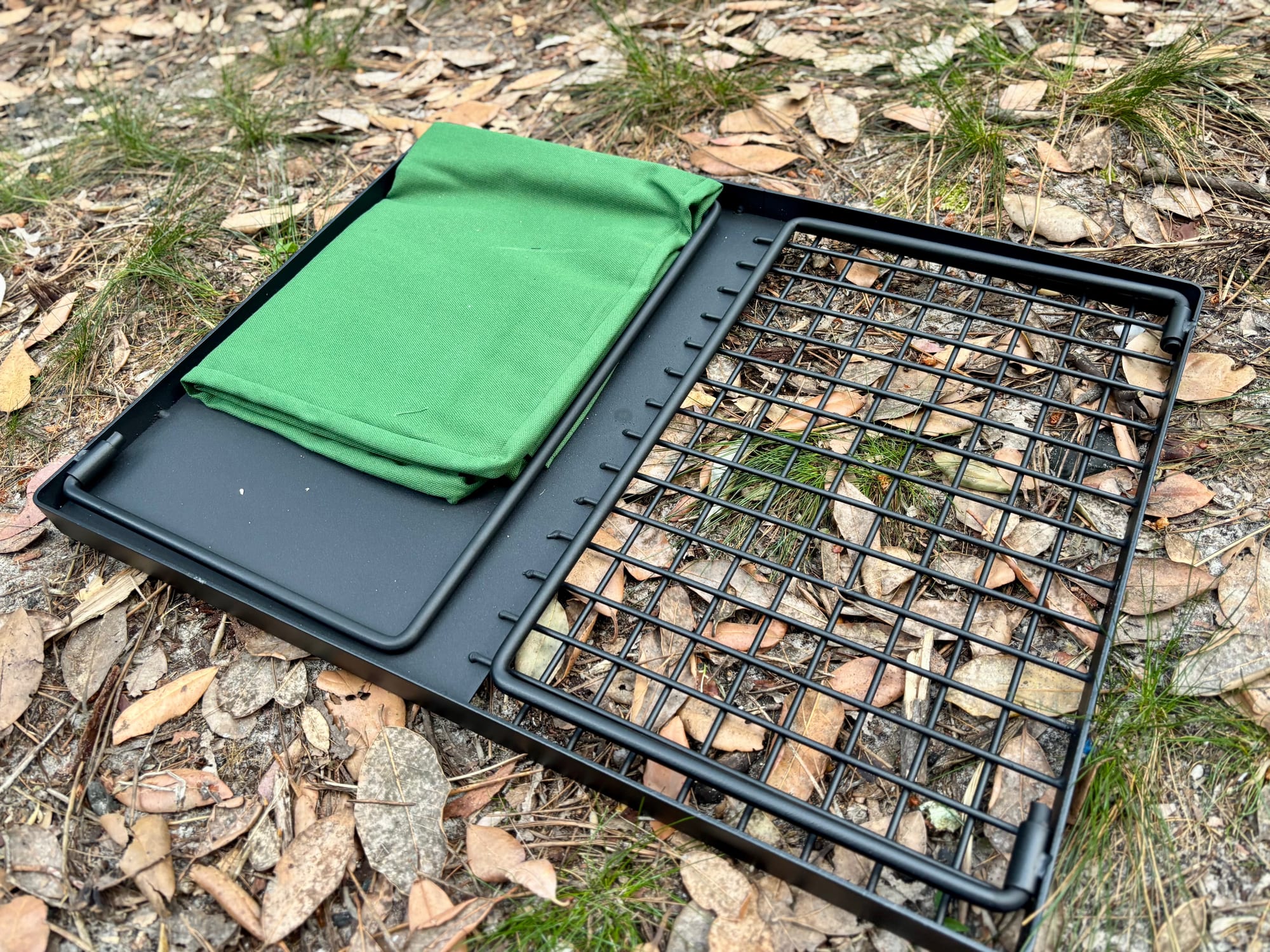 The Kings Camp Fire BBQ Plate with its legs folded, lying next to the green canvas bag