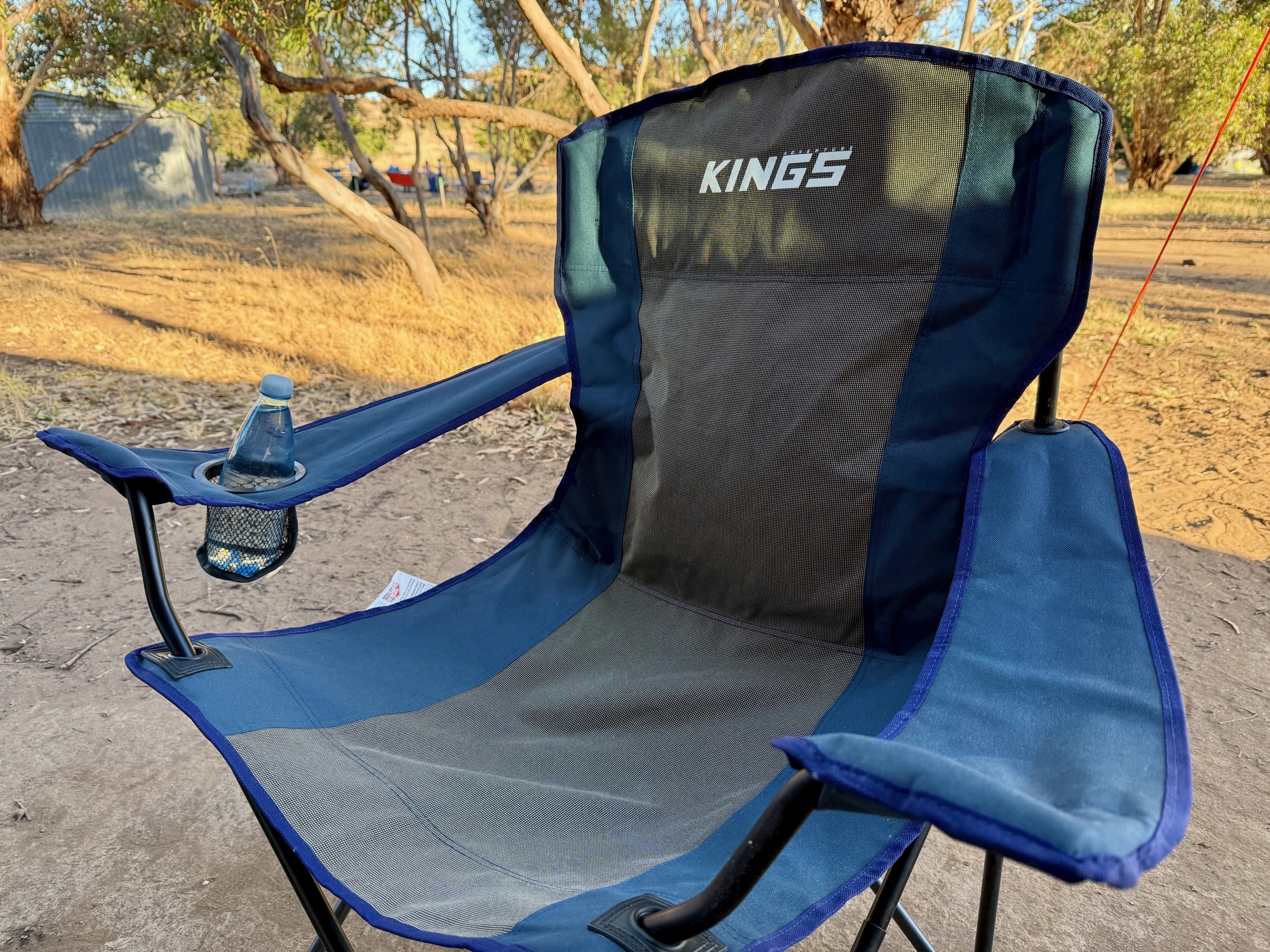 The Kings Essential Camping Chair at camp, with a water bottle in the armrest cup holder