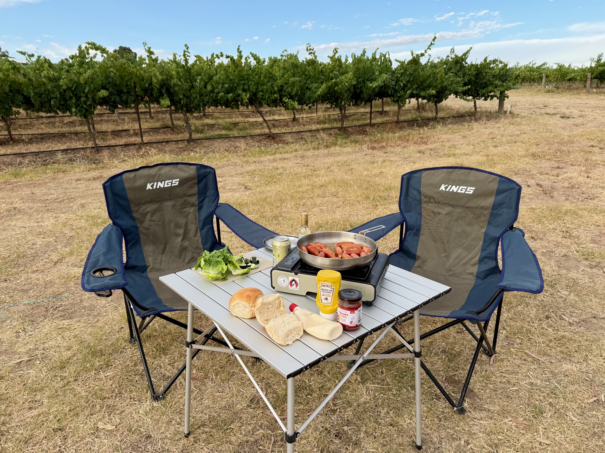 Two Kings Essential chairs at a camp table with food, next to a vineyard