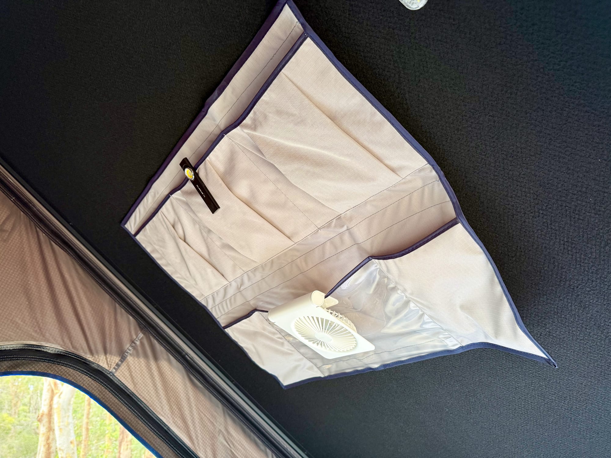 The storage pocket on the ceiling of a rooftop tent, with a small white fan in the clear pocket and a torch clipped to the top