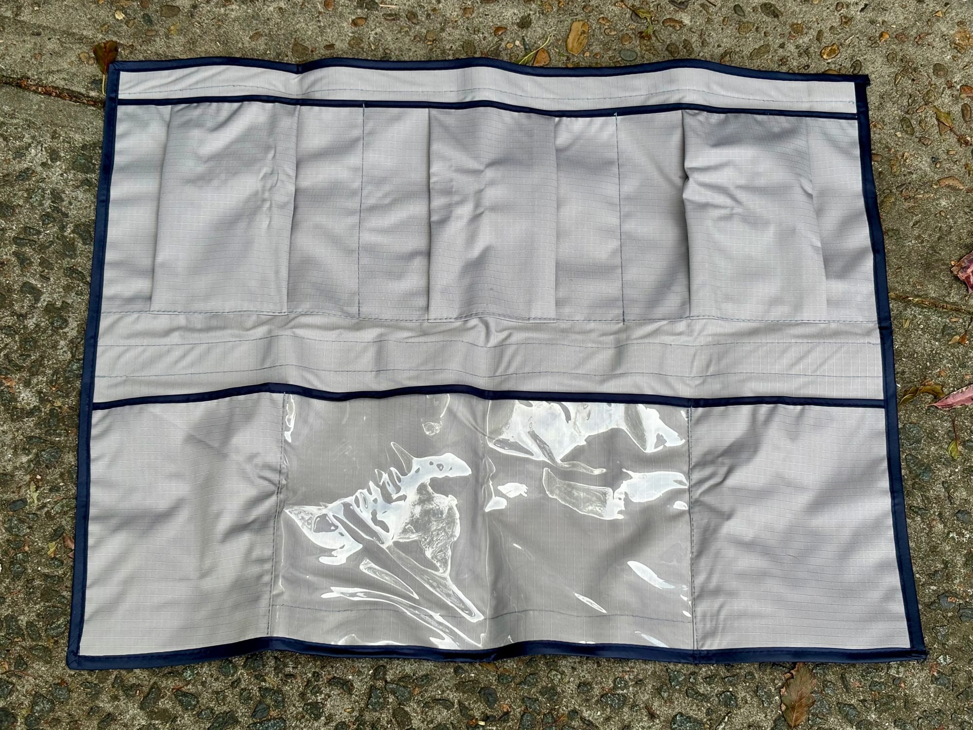 The grey storage pocket laid flat on concrete, showing the row of pockets at the top and the clear pocket at the bottom