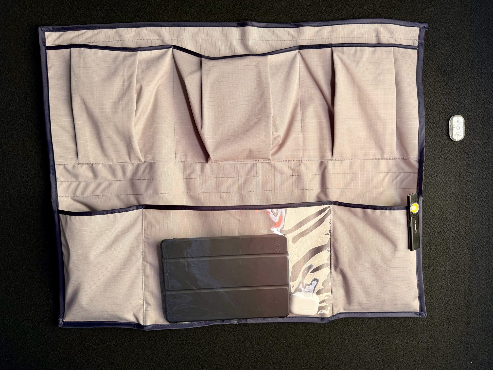 The storage pocket on the black carpeted ceiling of the tent, with a tablet and earbuds in the clear pocket and a torch clipped to the edge