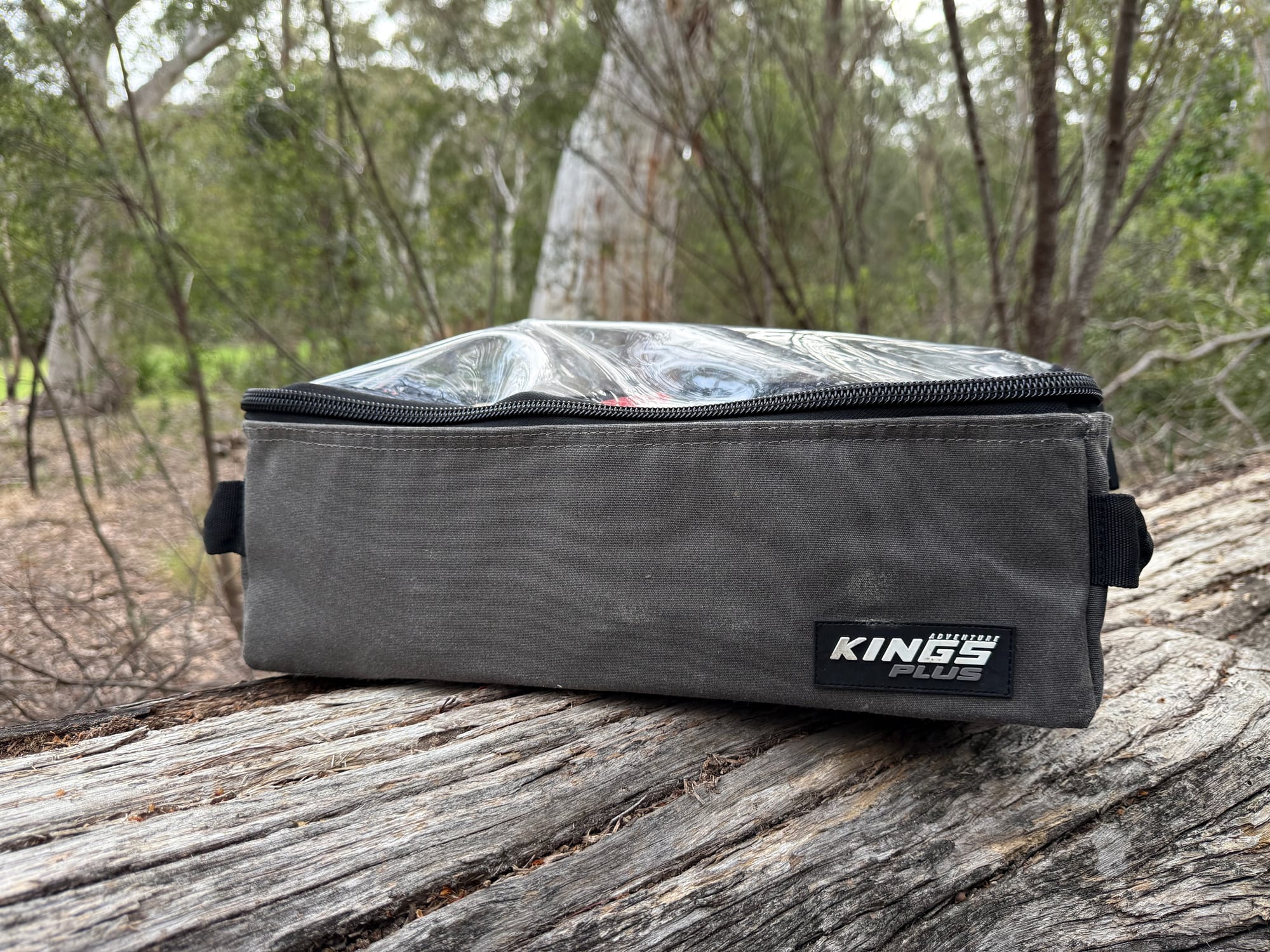 A grey Kings Plus clear top bag with its zipped clear lid on a log in the bush