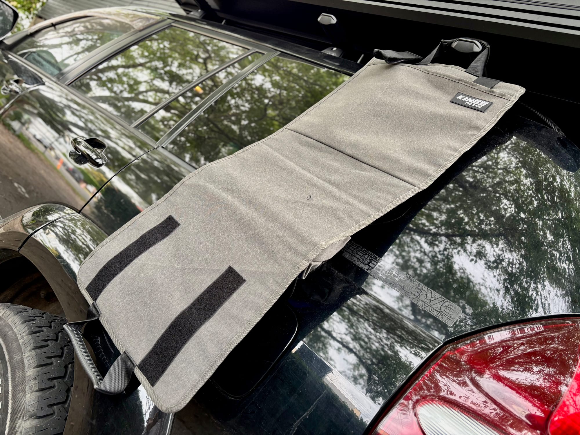 The grey canvas back of the Kings Plus Hanging Camp Caddy hanging from the roof rack down the side of a 4WD, with two hook and loop straps at the bottom