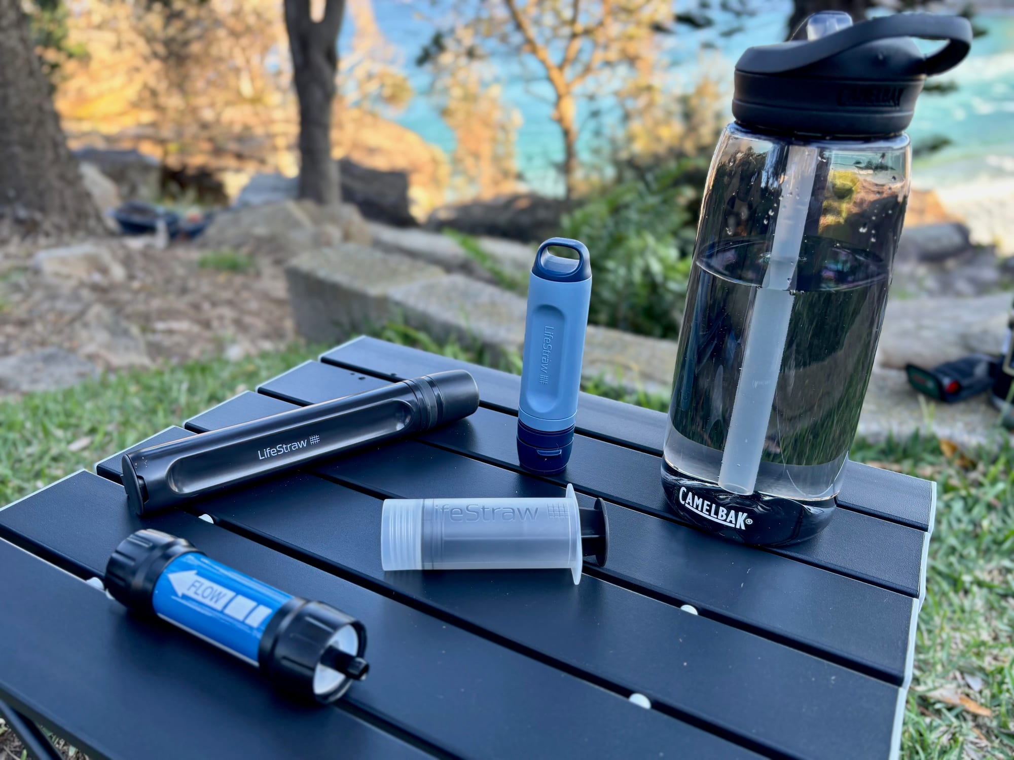 The Peak Solo, the LifeStraw Peak Series straw, the Sawyer Mini and the backwash syringe on a camp table, with a CamelBak bottle behind