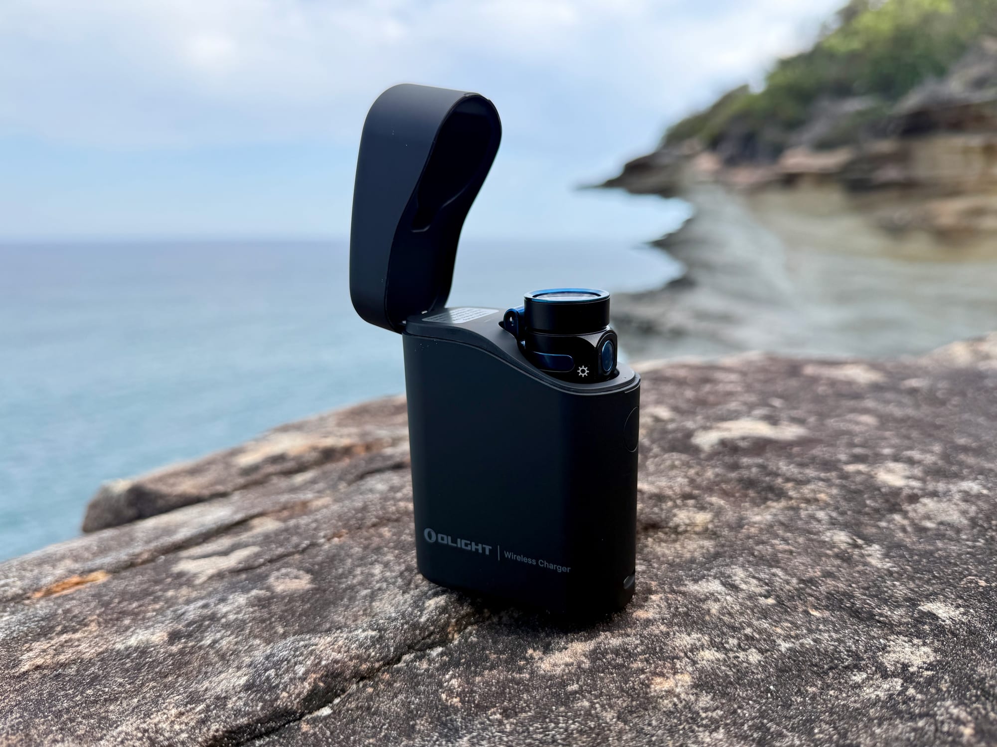 The Olight Baton 4 standing in its charging case with the lid open, on a rock above the sea